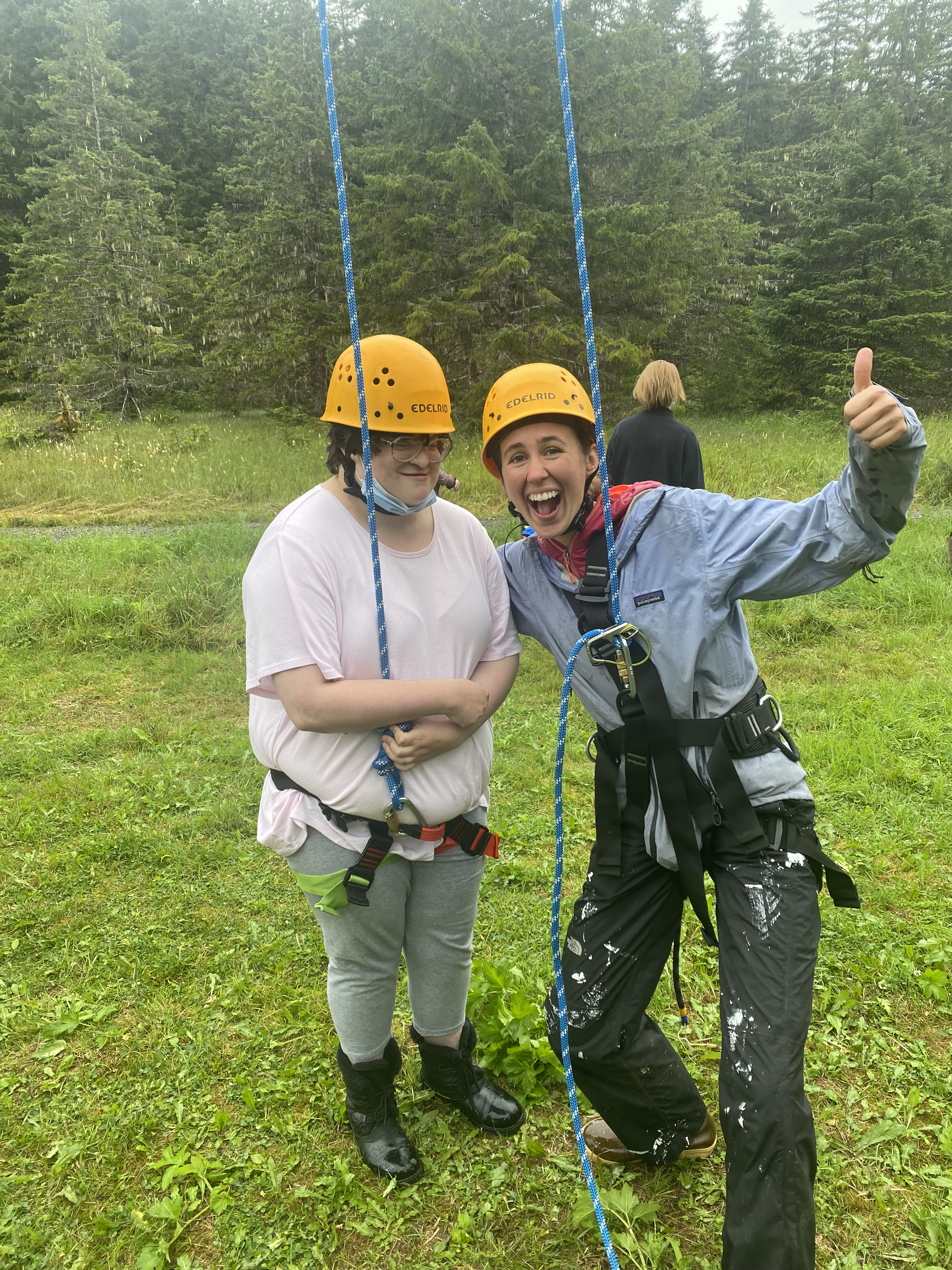 a young woman in white standing next to a young woman dressed in rain gear giving a thumbs up, both wearing climbing harnesses and roped up