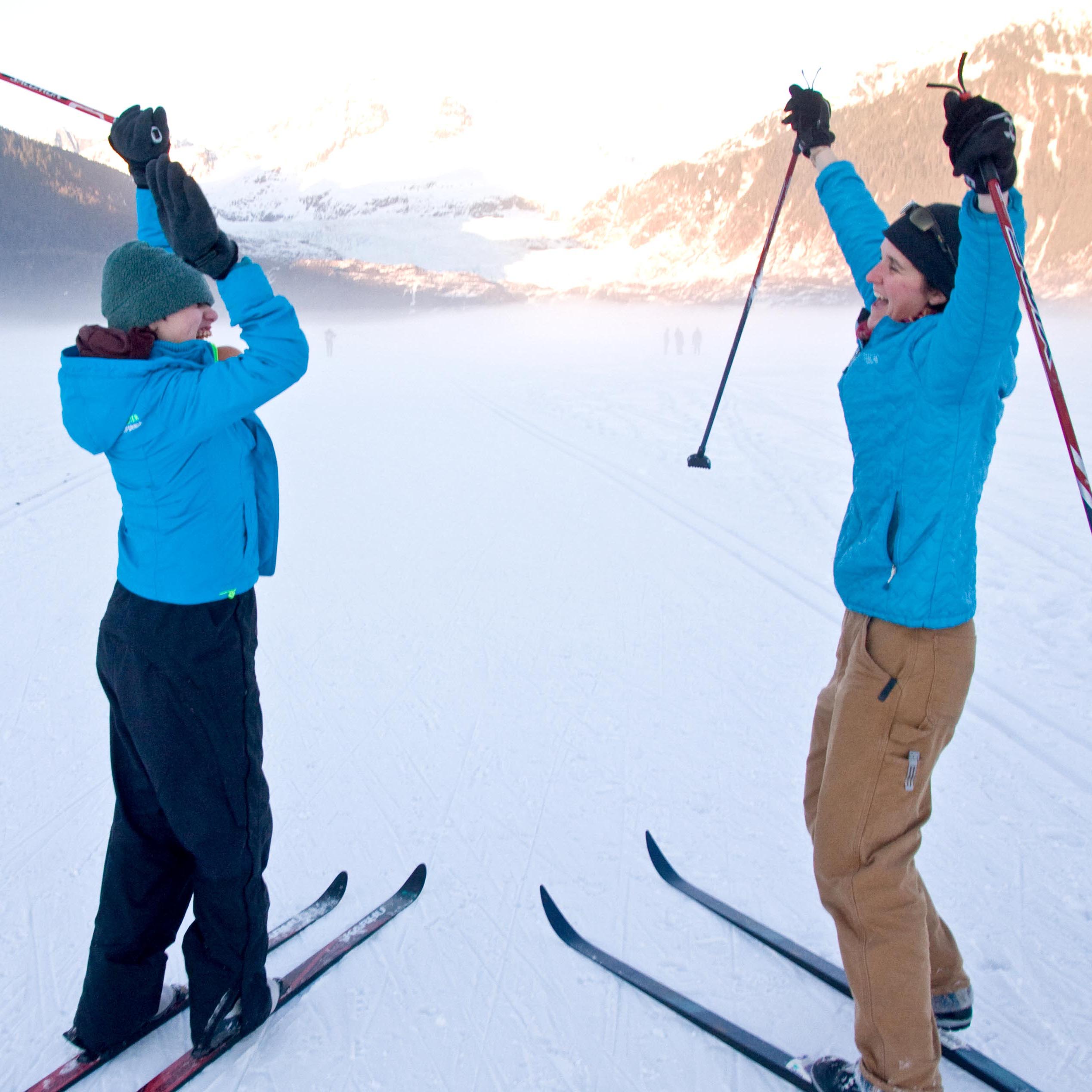 two women in bright blue jackets on cross country skis facing each other with their hands and poles in the air looking excited