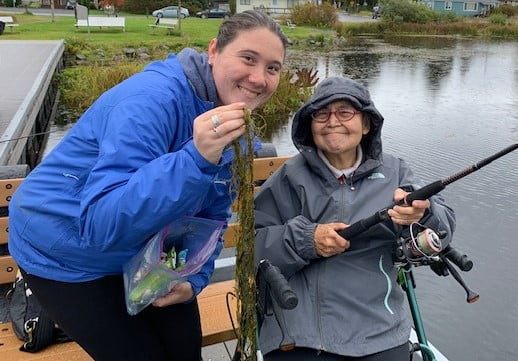 Anna wears a blue rain jacket and is holding a long string of pond weeds next to a senior outing participant holding a fishing rod.