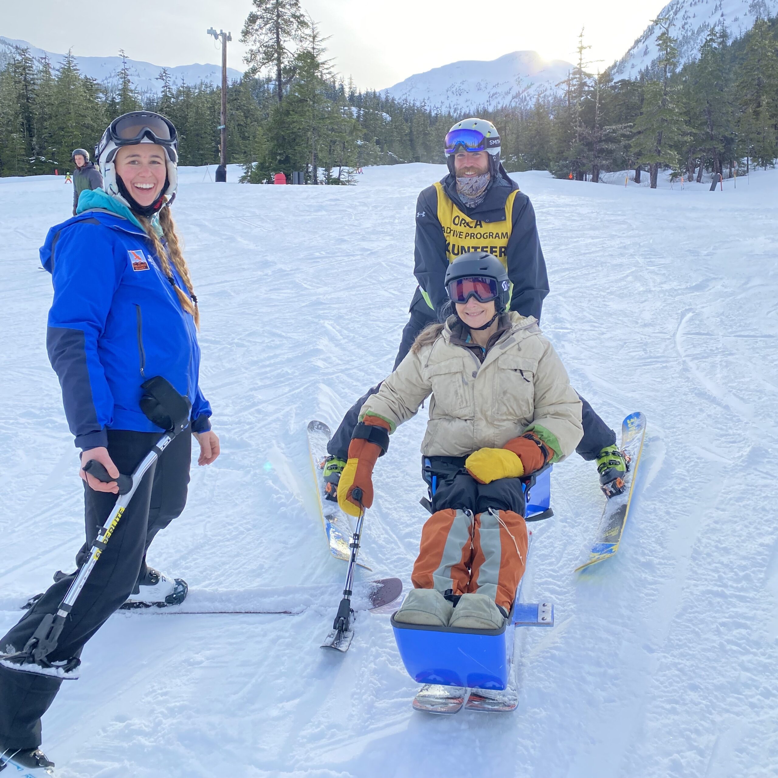 Woman learning to sit-ski poses for the camera with her instructors at the bottom of the porcupine slope.