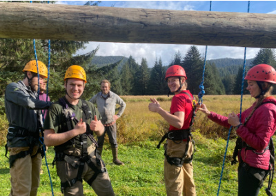 4 people in climbing helmets stand under a horizontal telephone pole, They are wearing climbing harnesses, with two people attached to climbing ropes. One man is giving a double thumbs up!