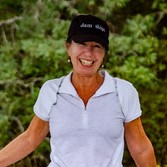 Board member Joan Sizemore is wearing a white polo shirt and navy blue ballcap. She is smiling in front of greenery.