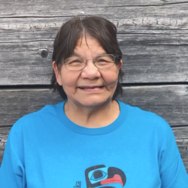 Board member Harriet Silva is smiling in front of a weather wood wall. Harriet has short black hair and wears wire rimmed glasses. She's wearing a turquoise t shirt with a formline eagle.