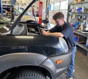 A young man is working in an auto shop. He leaning over the engine of a black vehicle.