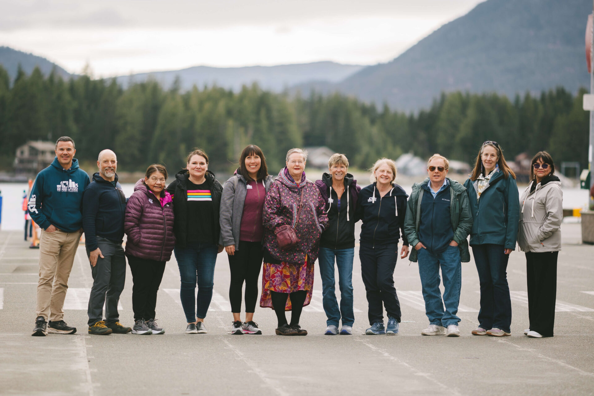 Group of eleven people standing together outdoors near the waterfront with forested mountains in the background, smiling and posing for a group photo on an overcast day.